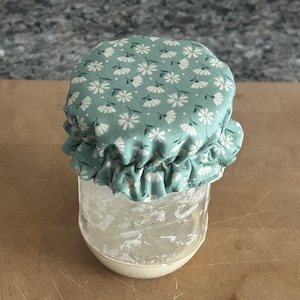 May include: A clear glass jar topped with a sage green fabric cover featuring a white daisy floral pattern. The fabric cover has a ruffled edge and is secured around the rim of the jar, creating a decorative seal for the container opening.