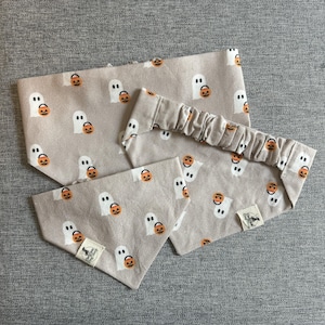Op de afbeelding: Een set Halloween-accessoires, waaronder een hoofdband en twee bandana's. De items zijn beige en hebben een herhaald patroon van witte geesten en oranje pompoenen. Elk item heeft een klein label met een vleermuislogo.