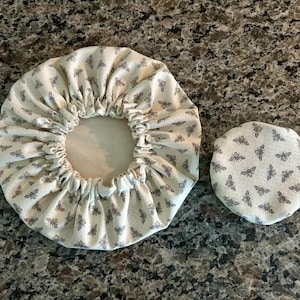 May include: Two circular fabric bowl covers featuring a repeating pattern of small grey bees on an off-white background. The larger cover has an elasticated gathered edge, while the smaller cover is a flat, circular piece of fabric.