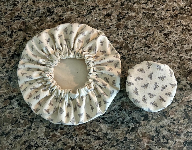 May include: Two circular fabric bowl covers featuring a repeating pattern of small grey bees on an off-white background. The larger cover has an elasticated gathered edge, while the smaller cover is a flat, circular piece of fabric.