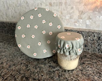 May include: A sage green fabric bowl cover and a matching ruffled jar cover, both featuring a repeating pattern of small white daisies with yellow centers. The jar cover is fitted over a glass jar containing a light-colored substance.