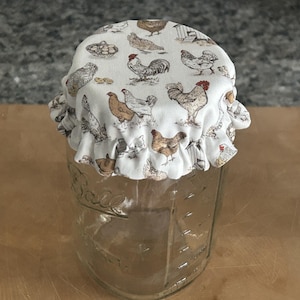 May include: A clear glass mason jar topped with a fabric cover featuring a repeating pattern of brown, white, and grey chickens, roosters, and small coops. The fabric has a ruffled edge that fits snugly over the rim of the jar.