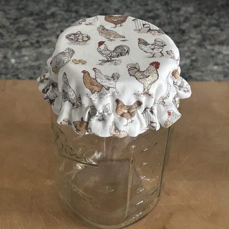 May include: A clear glass mason jar topped with a fabric cover featuring a repeating pattern of brown, white, and grey chickens, roosters, and small coops. The fabric has a ruffled edge that fits snugly over the rim of the jar.