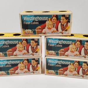 May include: A stack of five vintage Westinghouse Flash Cubes boxes. Each box features the text "Westinghouse Flash Cubes" and "3/12 Flashes" with a picture of children. The boxes are yellow and blue.
