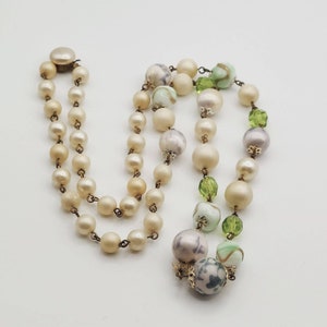 May include: A vintage multi-strand necklace with white and green glass beads. The necklace features a mix of round and faceted beads, with some beads having a swirl pattern.