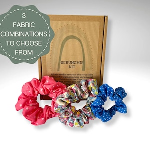 Könnte beinhalten: Ein Scrunchie-Kit mit drei Stoffkombinationen. Das Kit enthält eine braune Box mit dem Text "SCRUNCHIE KIT" und einem grünen Etikett mit der Aufschrift "3 STOFFKOMBINATIONEN ZUR AUSWAHL". Drei Scrunchies in Rosa, Blumenmuster und Blau mit weißen Punkten liegen vor der Box.