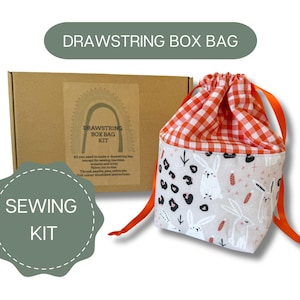 May include: A sewing kit for making a drawstring box bag. The kit includes fabric, thread, and instructions. The bag is made of a white fabric with a bunny print and a red and white gingham lining. The bag has a drawstring closure and orange cords.