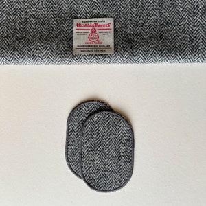 May include: Two grey oval-shaped coasters made of Harris Tweed fabric. The coasters have a black border and a herringbone pattern.