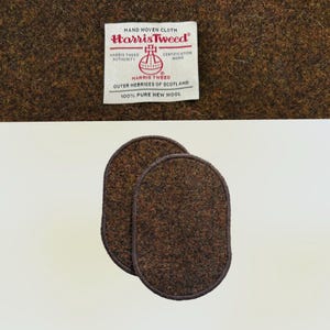 May include: Two oval brown wool patches with a dark brown border. The patches are made of Harris Tweed fabric. The Harris Tweed label is visible in the background, with text that reads "HAND WOVEN CLOTH Harris Tweed OUTER HEBRIDES OF SCOTLAND 100% PURE NEW WOOL."