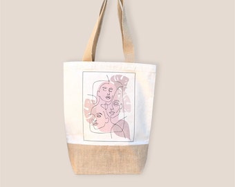 May include: A white tote bag with a brown burlap bottom. The bag features a graphic of three faces drawn in black lines with pink and purple watercolor washes. The faces are surrounded by leaves.