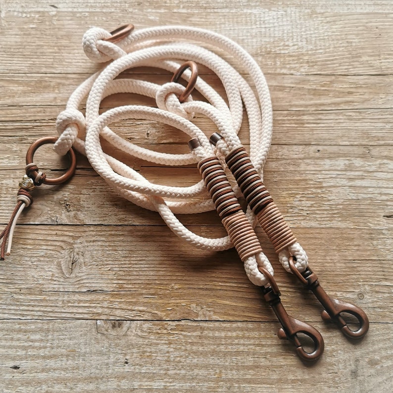 Pode incluir: Duas trelas de c&atilde;o de algod&atilde;o branco com detalhes em listras marrons e brancas e ferragens de cobre. As trelas est&atilde;o enroladas juntas e t&ecirc;m um anel em uma extremidade e um clipe na outra.