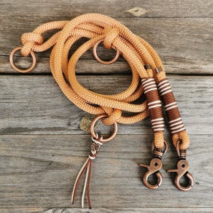 May include: A brown and white rope dog leash with two copper clips and a copper paw charm. The leash is coiled on a wooden surface.