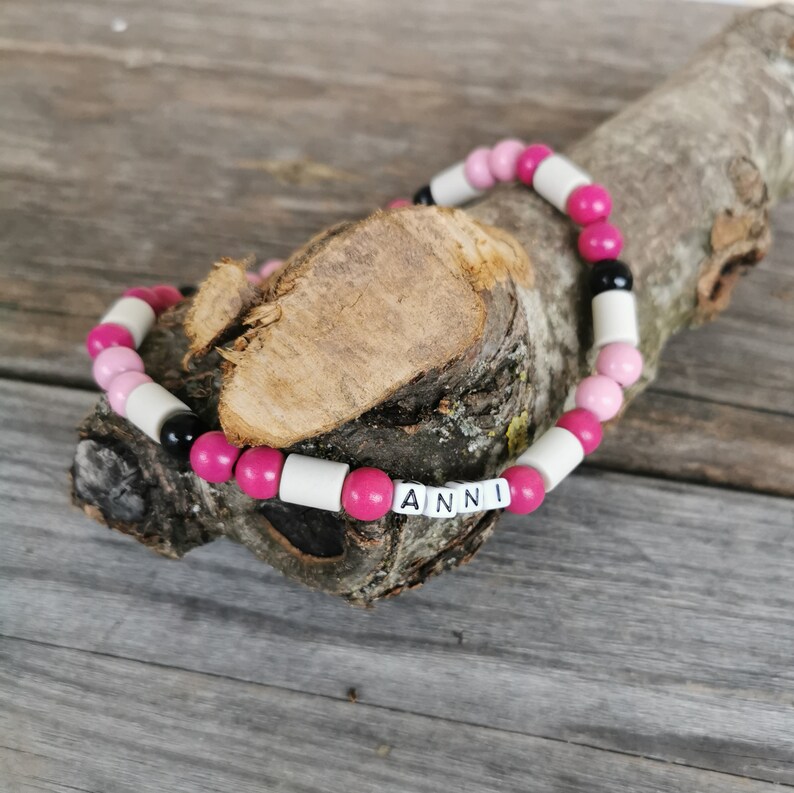 May include: A pink and white beaded bracelet with black beads and the name "ANNI" spelled out in white letters.