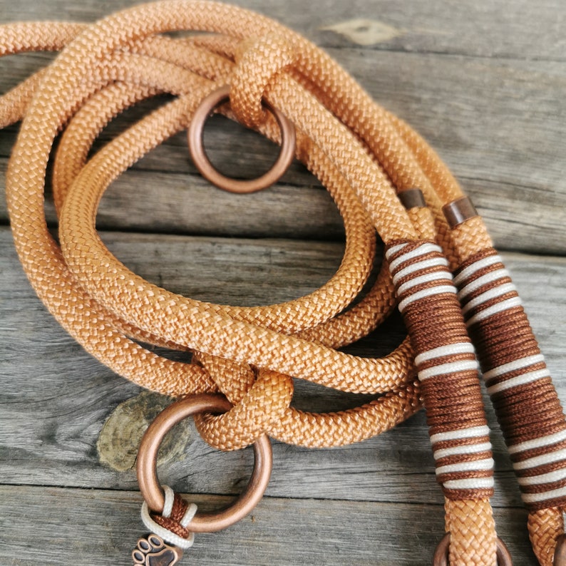 May include: A close-up of a brown and white rope dog leash with copper hardware. The leash is made of a thick, braided rope and has a copper ring at one end and a copper paw print charm at the other.