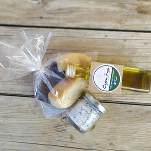 Puede incluir: Una cesta de regalo con pan, aceite de oliva y sal marina. El pan está en una bolsa de celofán transparente atada con una banda elástica dorada. También se incluye una pequeña botella de vidrio de aceite de oliva y un tarro de sal marina. La etiqueta de la botella de aceite de oliva dice "Cretan Farm".