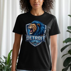 Op de afbeelding: Een zwart T-shirt met korte mouwen voorzien van een afbeelding van een brullende leeuw met blauwe manen in een zilveren schildembleem. Onder de leeuw staat het woord DETROIT in dikke zilveren letters boven een silhouet van een skyline.