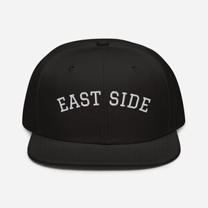 Può includere: Cappellino da baseball nero con visiera piatta, con la scritta bianca ricamata "EAST SIDE" sul davanti. Il cappellino ha una corona strutturata. Un classico stile di cappello.