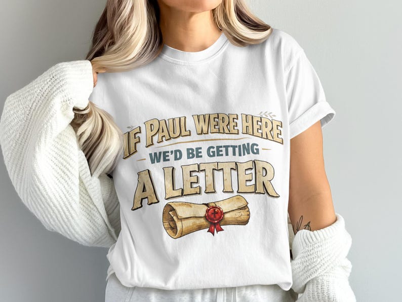 White t-shirt with text "IF PAUL WERE HERE WE'D BE GETTING A LETTER" and a scroll graphic.