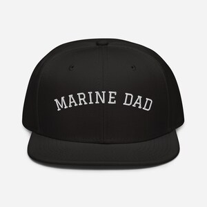 Könnte beinhalten: Schwarze Baseballkappe mit dem weißen Schriftzug "MARINE DAD" auf der Vorderseite. Die Kappe hat einen flachen Schirm und eine strukturierte Krone. Der Text ist fett und ohne Serifen. Ein modisches Accessoire.