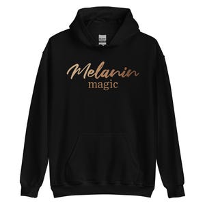 May include: Black hooded sweatshirt with the words "Melanin magic" in a copper-colored script across the chest. The sweatshirt has a front pocket and a hood with a drawstring.