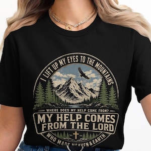 May include: A black short-sleeved t-shirt featuring a circular graphic with a mountain landscape, a flying eagle, and pine trees. The text reads: I LIFT UP MY EYES TO THE MOUNTAINS, WHERE DOES MY HELP COME FROM? MY HELP COMES FROM THE LORD, WHO MADE HEAVEN & EARTH.
