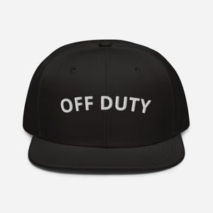 Peut inclure: Casquette de baseball noire avec une visière plate. Les mots "OFF DUTY" sont brodés en blanc sur le devant. La casquette a une coupe structurée et une visière incurvée.