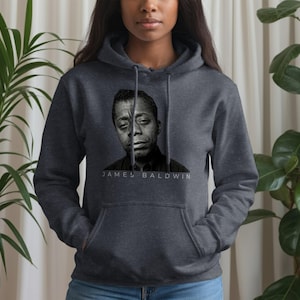 Könnte beinhalten: Ein dunkelgrauer Kapuzenpullover mit einem Schwarz-Weiß-Porträt von James Baldwin. Der Name "JAMES BALDWIN" ist darunter in Weiß gedruckt. Der Hoodie hat eine Kapuze, eine Vordertasche und lange Ärmel.