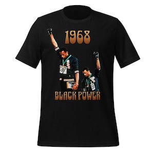 Puede incluir: Camiseta negra con un gráfico de dos atletas con los puños en alto. El texto "1968" está encima de los atletas, y "BLACK POWER" debajo. Los atletas llevan uniformes negros y verdes.