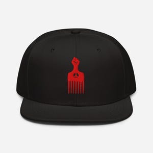May include: Black flat brimmed baseball cap with a red embroidered design of a raised fist holding a comb with a peace sign in the middle.