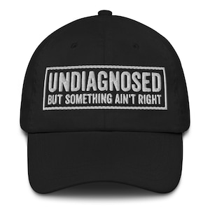 Könnte beinhalten: Schwarze Baseballkappe mit dem gestickten Text "UNDIAGNOSED BUT SOMETHING AIN'T RIGHT" in Weiß. Die Kappe hat einen gebogenen Schirm und einen Knopf oben. Ein lässiges Accessoire.