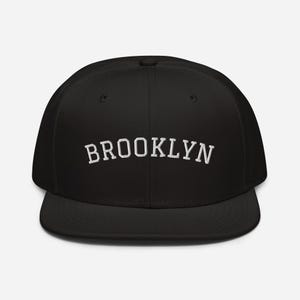 Può includere: Un cappellino da baseball nero con visiera piatta. La parola "BROOKLYN" è ricamata in lettere bianche sulla parte anteriore. Il cappellino ha una vestibilità strutturata e una visiera curva.