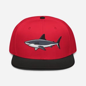 Puede incluir: Gorra de béisbol roja con visera negra, con un diseño de tiburón bordado. El tiburón está representado en tonos grises y blancos, con un contorno detallado. La gorra tiene una visera plana clásica.