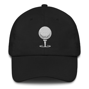 Può includere: Cappellino da baseball nero con un design a palla da golf bianca ricamata. La pallina da golf è su un tee. Il cappellino ha una visiera curva e un cinturino regolabile. Un accessorio per i golfisti.