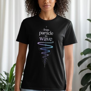 Peut inclure: Un t-shirt noir à manches courtes avec le texte blanc "from particle to wave" centré sur la poitrine. Sous le texte se trouve un graphique représentant une traînée lumineuse tourbillonnante dans des tons de violet et de bleu.