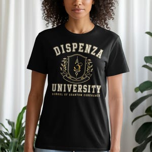 May include: A black short-sleeve t-shirt featuring a gold and white graphic print. The design includes the text DISPENZA UNIVERSITY, a central crest with a symbol, and the text SCHOOL OF QUANTUM COHERENCE printed in a collegiate style font.