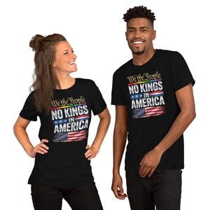 Può includere: T-shirt nere con la scritta "We the People NO KINGS IN AMERICA" in bianco, blu e rosso. Il design include una bandiera americana. Le magliette sono a maniche corte e indossate da due persone.