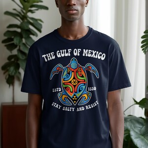 May include: A navy blue t-shirt featuring a colorful graphic of a sea turtle with intricate patterns in red, orange, green, and blue. The text reads THE GULF OF MEXICO, ESTD 1550, and STAY SALTY AND RESIST in white lettering on the front.