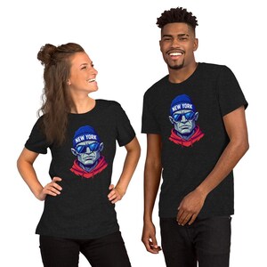 Op de afbeelding: Twee mensen dragen donkergrijze t-shirts. Elk shirt heeft een afbeelding van een man met een blauwe muts met "NEW YORK" erop, een zonnebril en een rode hoodie. De afbeelding is een gestileerde illustratie.