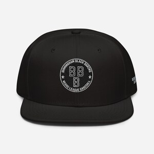 Puede incluir: Gorra de béisbol negra con el logotipo blanco de los Birmingham Black Barons. El logotipo es un diseño circular con el texto "Birmingham Black Barons Negro League Baseball" alrededor de las letras "BB B". La gorra tiene una visera plana.