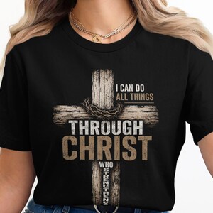 May include: A black short-sleeved t-shirt featuring a graphic of a wooden cross with a crown of thorns. The text reads I CAN DO ALL THINGS THROUGH CHRIST WHO STRENGTHENS ME in a mix of white and tan distressed lettering centered on the chest.