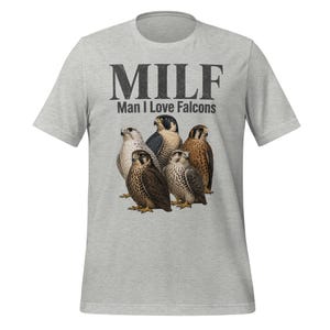 Può includere: T-shirt grigio chiaro con una grafica di cinque falchi in varie tonalità di marrone e bianco. Sopra gli uccelli, il testo "MILF" e sotto, "Man I Love Falcons" in un font invecchiato.