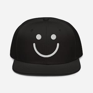 Könnte beinhalten: Schwarze Baseballkappe mit einem weißen, gestickten Smiley. Die Kappe hat einen flachen Schirm, eine strukturierte Vorderseite und einen verstellbaren Snapback-Verschluss. Dies ist ein lässiges Accessoire.