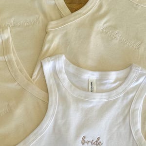 May include: Four sleeveless tank tops in cream and white. Three cream-colored tops are embroidered with the word "bridesmaid" in a cursive font. A white tank top is embroidered with the word "bride" in a similar font.
