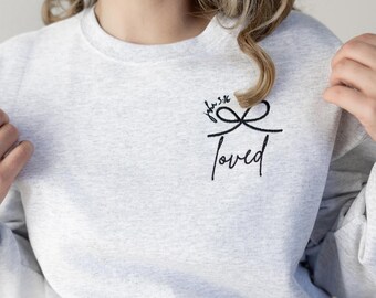 Può includere: Una felpa girocollo grigio chiaro con una grafica nera sulla parte superiore sinistra del petto. Il design include un simbolo dell'infinito stilizzato, il testo "John 3:16" in caratteri piccoli e la parola "loved" in un carattere corsivo più grande.