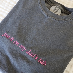 以下が含まれることがあります： ピンク色の刺繍で「put it on my dad's tab」と書かれたチャコールグレーのTシャツ。折りたたまれたシャツは、白くテクスチャ加工された表面に置かれています。クルーネックで、Comfort Colorsのタグが付いています。