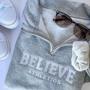Puede incluir: Sudadera gris con cremallera blanca y el texto "BELIEVE ATHLETICS" en blanco. Un par de gafas de sol marrones están encima de la sudadera.