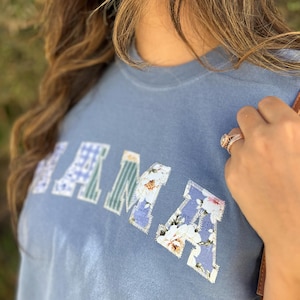 以下が含まれることがあります： パッチワーク文字で「MAMA」と書かれた青いTシャツ。文字は花柄やチェック柄で装飾されています。このシャツは、リラックスしたカジュアルなスタイルです。