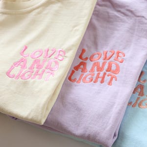 May include: Four t-shirts in different colors with the phrase "LOVE AND LIGHT" embroidered in pink on the front of each shirt.