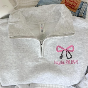 May include: A gray sweatshirt with a zipper and a pink bow with a stethoscope and the text "Kayla, RN, BSN" embroidered on the front.