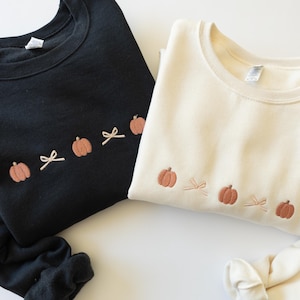 May include: A black sweatshirt and a cream-colored sweatshirt, each featuring an embroidered horizontal row of small orange pumpkins alternating with light beige bows across the chest area. Both garments have long sleeves and a crew neckline.